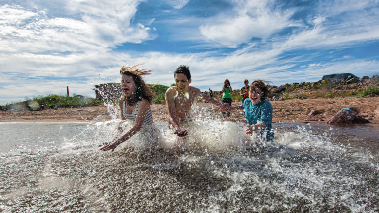 Kolme lasta juoksee veteen hiekkaiselta uimarannalta. Kaksi henkilöä seisoo taustalla. Taivas pilvinen mutta aurinko paistaa.