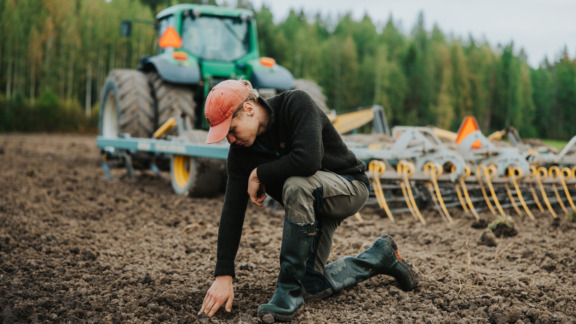 Viljelijä kumartuneena koskettamaan maata punaisen traktorin edessä pellolla.