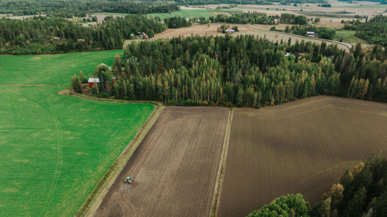 Ilmakuva. Peltomaata ja metsää. Pellolla traktori.