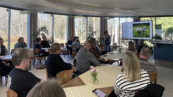 Kuva VERKKO-tapahtumasta Kymen Paviljongissa. Etualalla osallistujia pöytien ääressä. Taka-alalla puhuja esittelemässä uudistavaa viljelyä. Diaestityksessä kuva lehmästä laitumella.